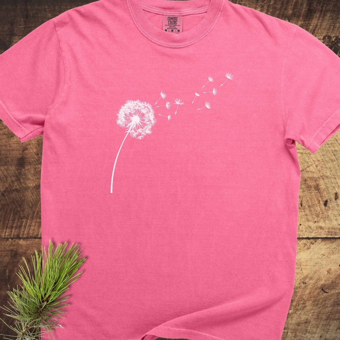 Dandelion Comfort Colors 1717 Tee, pink, featuring a white dandelion print, laid flat on wood. Made from soft-washed, garment-dyed 100% ring-spun cotton.
