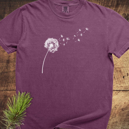 Dandelion Comfort Colors 1717 Tee in plum, short-sleeved, with a white dandelion graphic on chest, laid flat on wood, pine branch nearby.