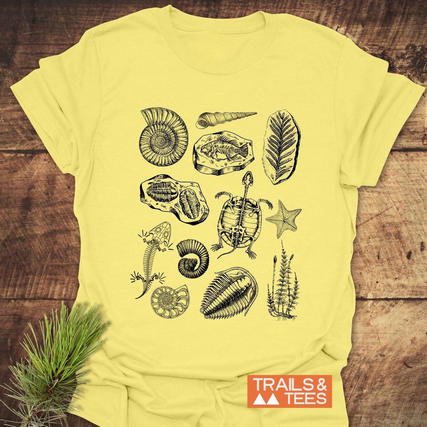 Fossile Vintage Tattoo T-Shirt featuring black line-drawn fossil illustrations, including ammonite, trilobites, and turtle skeleton, displayed on a rustic wooden surface with TRAILS & TEES logo.