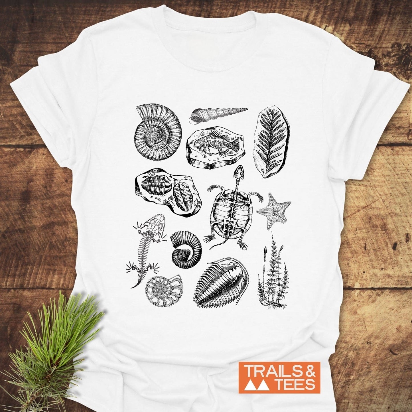 Fossile Vintage Tattoo T-Shirt featuring black line-drawn fossils and prehistoric specimens, including ammonite shells and trilobites, with TRAILS & TEES logo on a wooden surface.