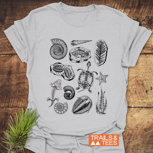 Fossile Vintage Tattoo T-Shirt featuring hand-drawn fossils and skeletons, including ammonites, trilobites, and ancient plants, with TRAILS & TEES logo on a wooden surface.