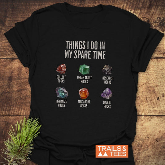 Sparetime Rock Lover T-Shirt featuring playful rock-themed illustrations and captions, displayed on a wooden surface with a TRAILS & TEES logo visible.