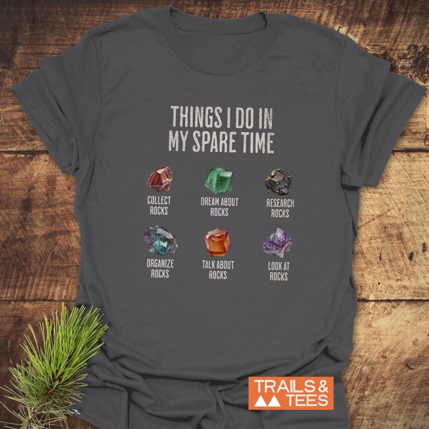 Sparetime Rock Lover T-Shirt featuring six rock-related pastimes graphic, laid on a wooden surface, with “TRAILS & TEES” logo in the corner.