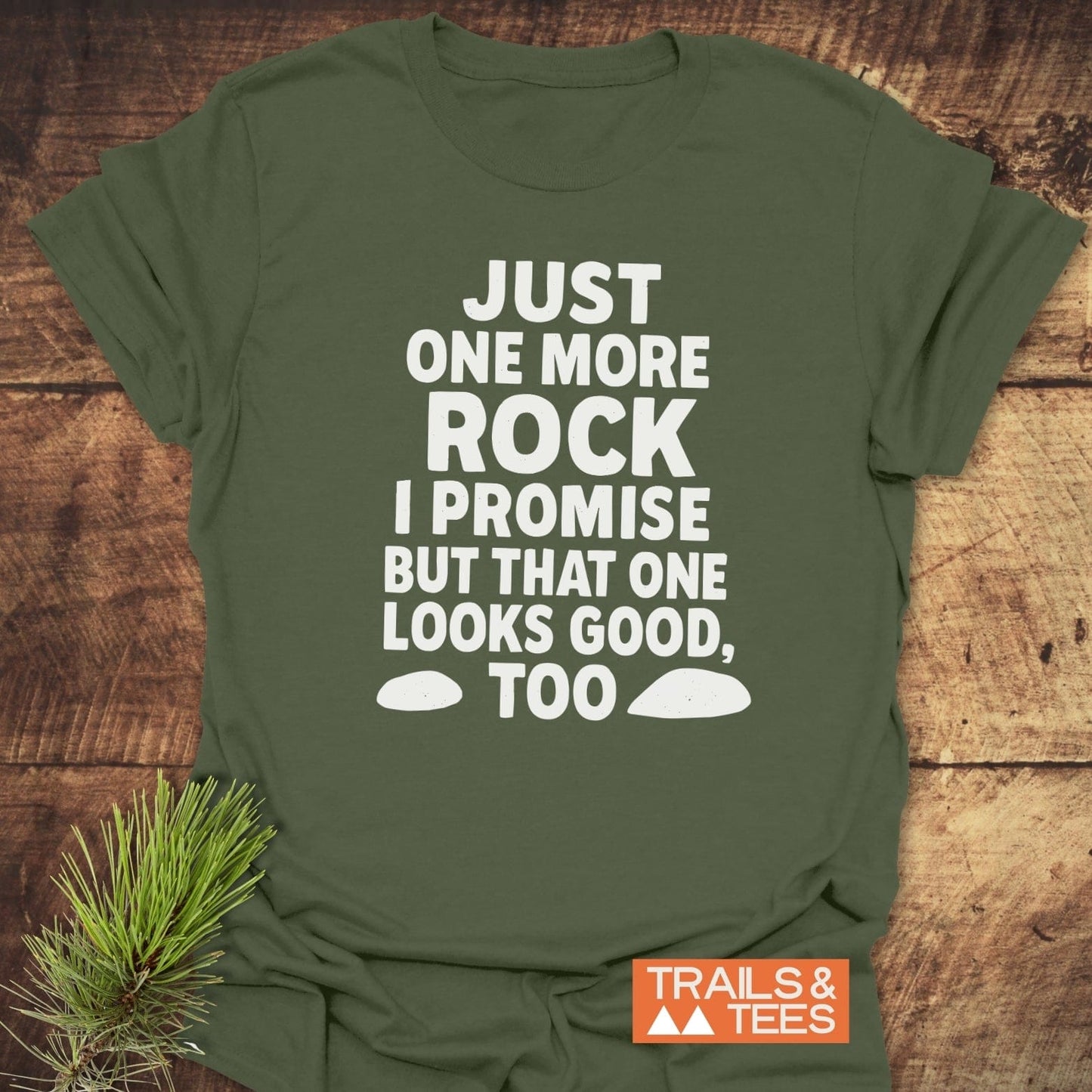 Forest-green T-shirt with bold white text: JUST ONE MORE ROCK I PROMISE BUT THAT ONE LOOKS GOOD, TOO, featuring rock silhouettes. TRAILS & TEES logo included.