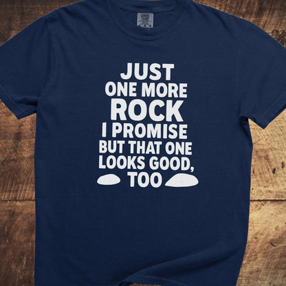 Just One More Rock Funny Comfort Colors 1717 Tee, navy-blue, with bold white slogan and rock shapes, laid flat on a wooden surface.