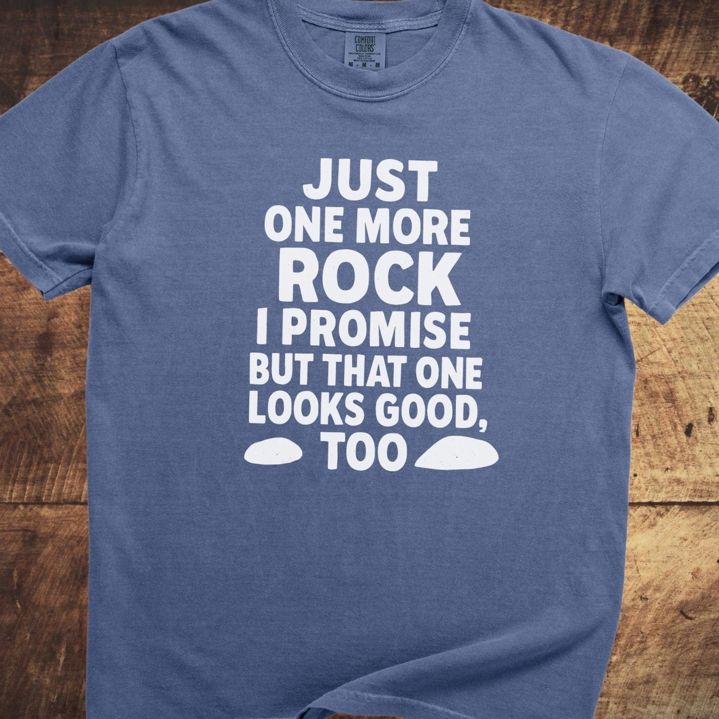 Just One More Rock Funny Comfort Colors 1717 Tee, blue with playful white text and rock graphics, laid flat on a wooden surface.