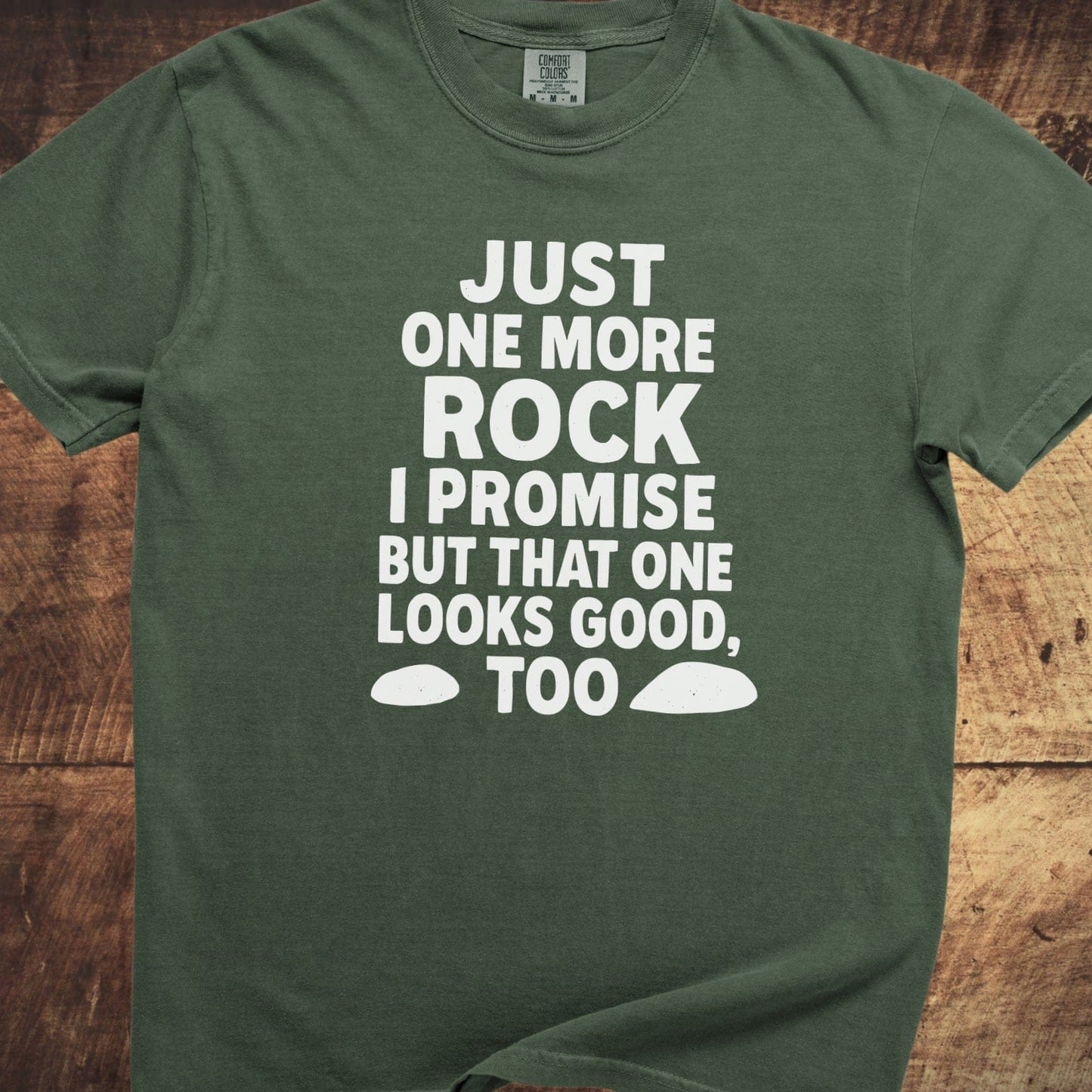 Just One More Rock Funny Comfort Colors 1717 Tee, dark-green, short-sleeve, with bold white slogan and rock shapes, laid flat on a wooden surface.
