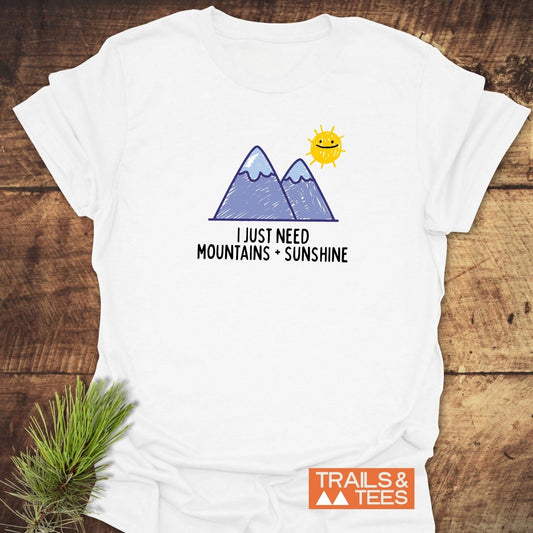 Mountains+Sunshine T-Shirt featuring a hand-drawn graphic of purple mountains and a smiling sun, with the slogan “I JUST NEED MOUNTAINS + SUNSHINE.”