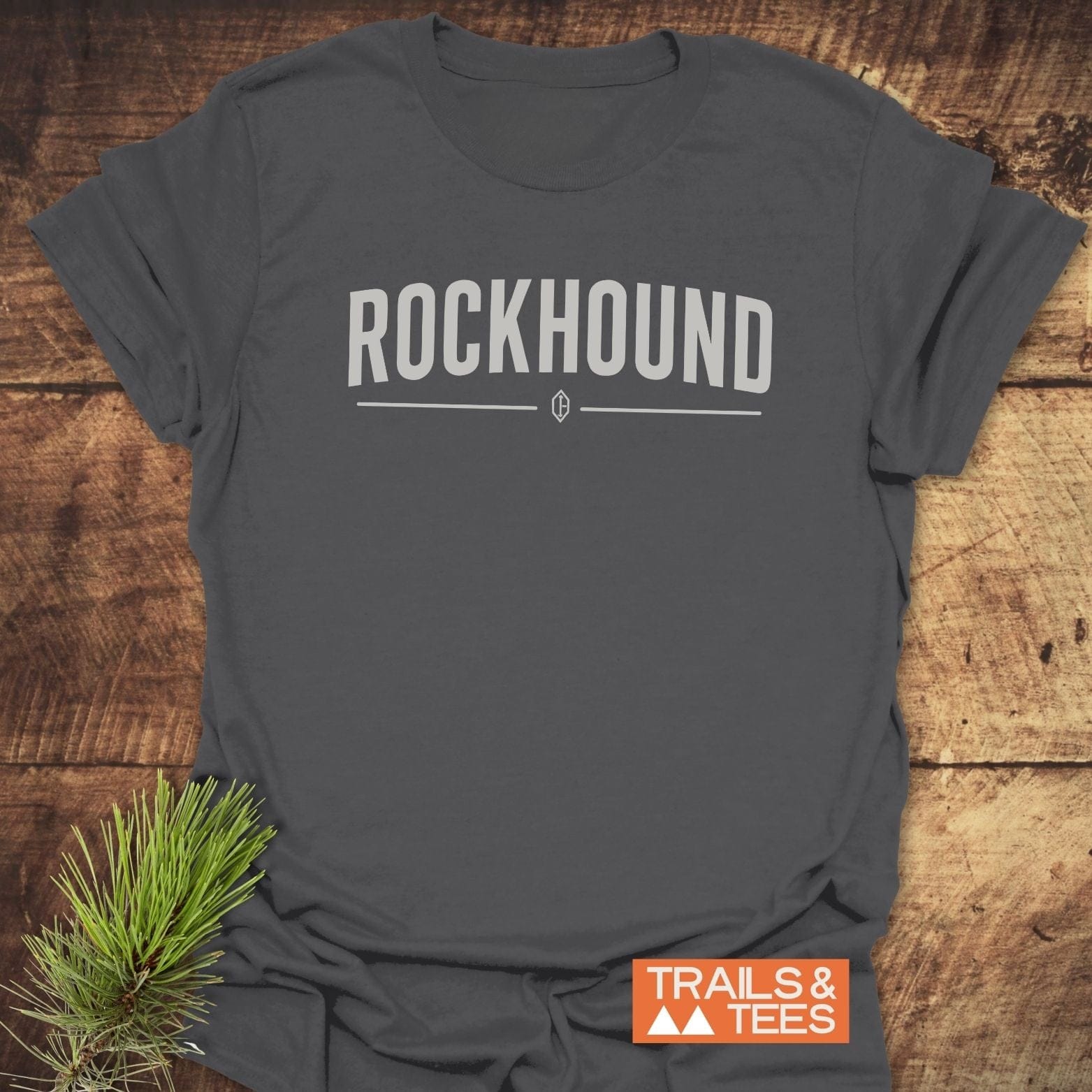 Charcoal-gray T-shirt with ROCKHOUND in bold letters, gem icon below. Features TRAILS & TEES logo and pine twig accent. Rockhound Rock Lovers T-Shirt.
