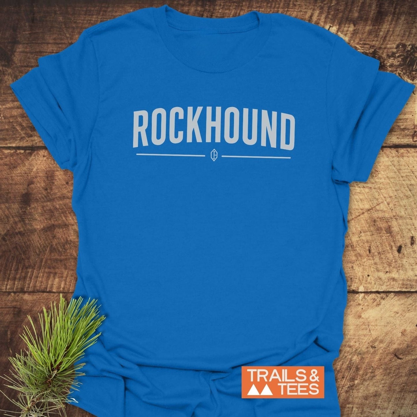 Bright blue Rockhound Rock Lovers T-Shirt on wood surface, featuring bold white lettering and emblem, with Trails & Tees logo and a pine sprig accent.
