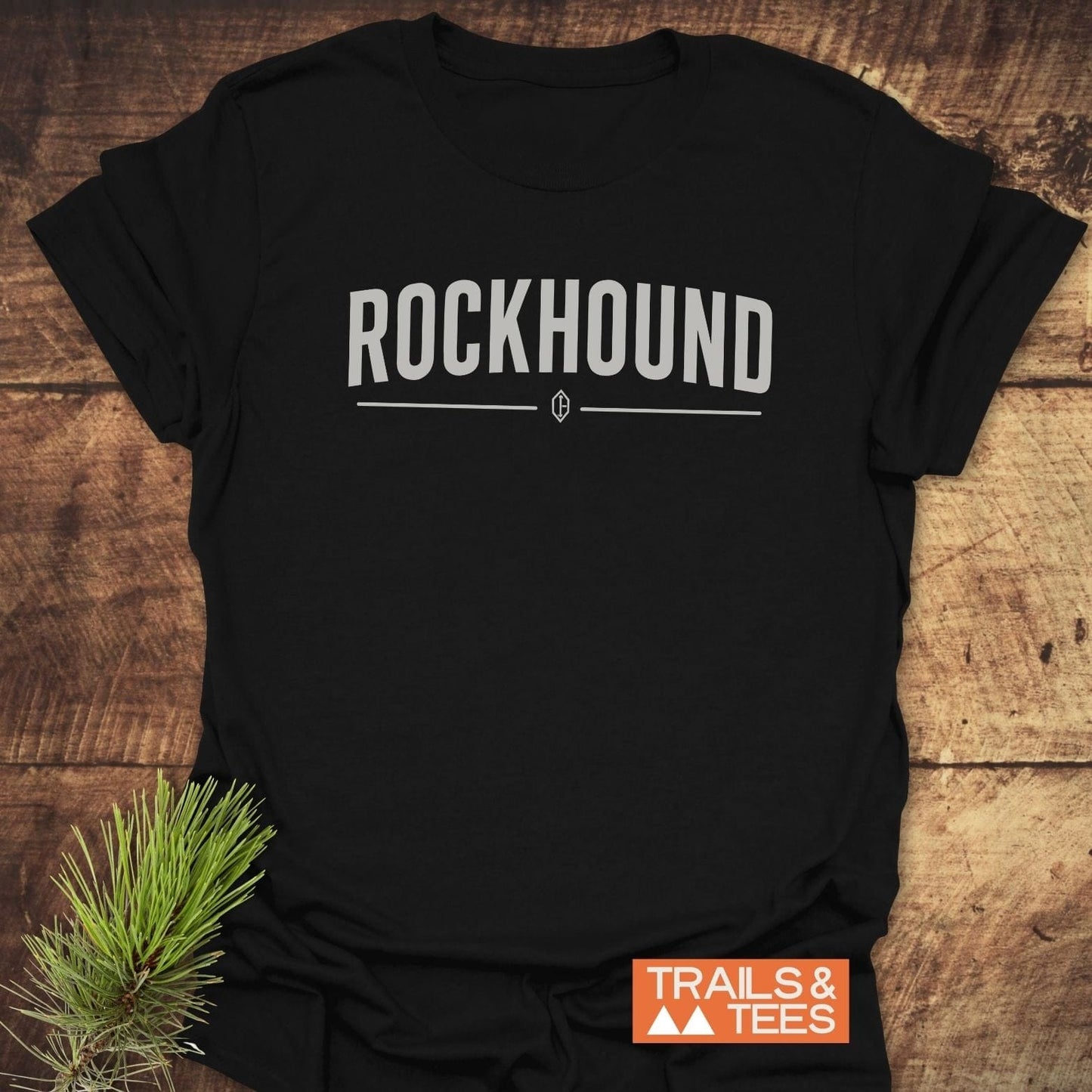 Flat-lay of the Rockhound Rock Lovers T-Shirt with ROCKHOUND text, crystal icon, and Trails & Tees logo, complemented by a pine sprig.