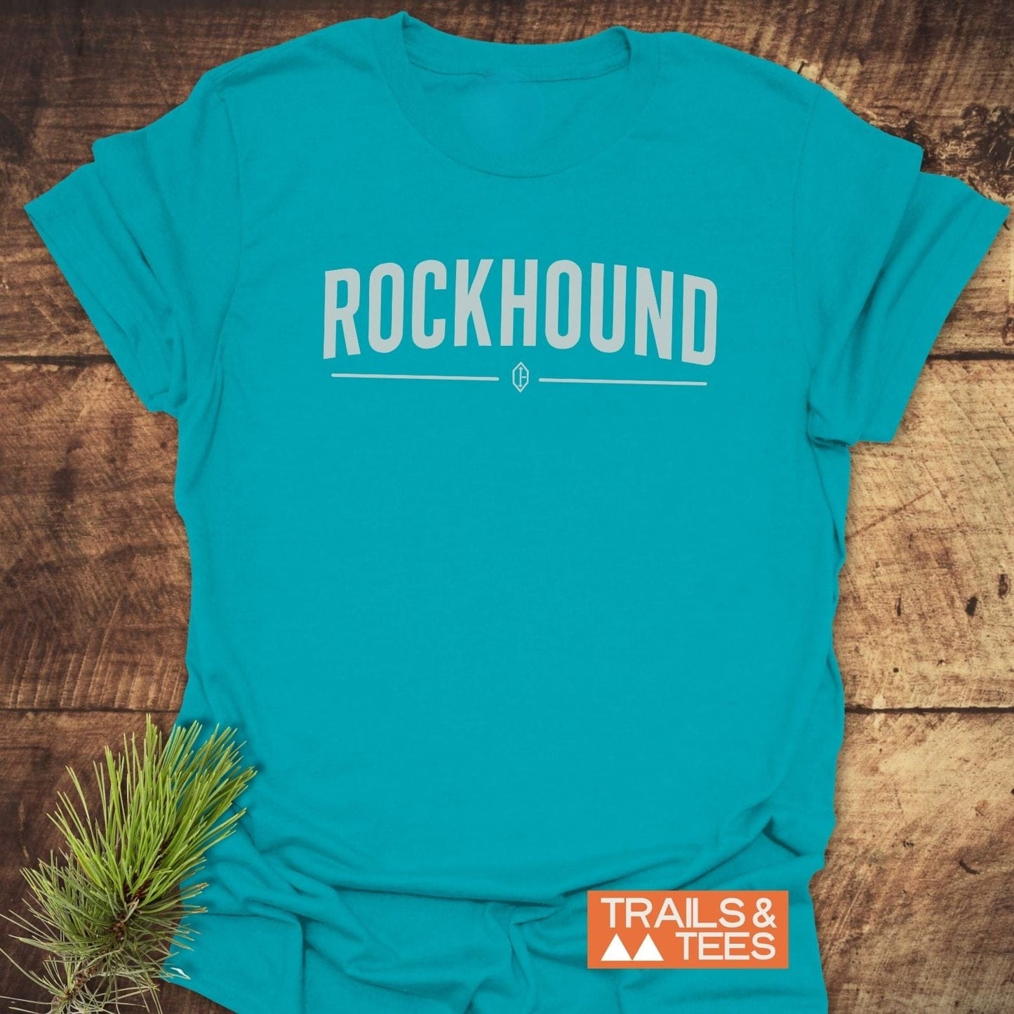 Rockhound Rock Lovers T-Shirt with bold ROCKHOUND text, gem icon, and Trails & Tees logo, displayed on wood with a pine sprig.