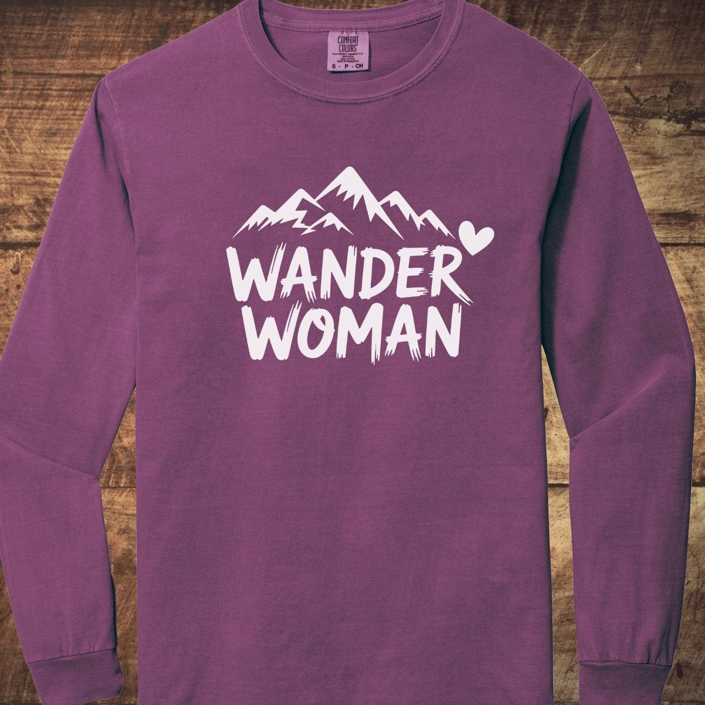 Wander Woman Comfort Colors Long-Sleeve Tee with a white mountain graphic and WANDER WOMAN text on the front, laid on a wooden surface.