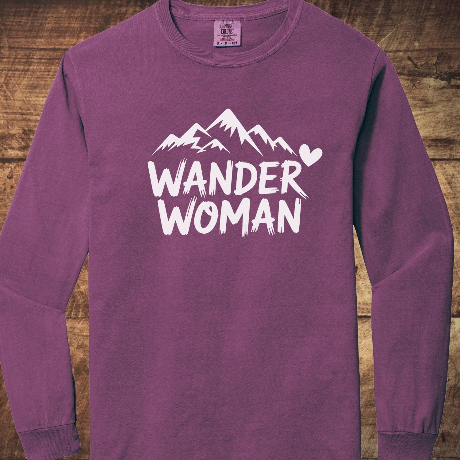 Wander Woman Comfort Colors Long-Sleeve Tee with a white mountain graphic and WANDER WOMAN text on the front, laid on a wooden surface.