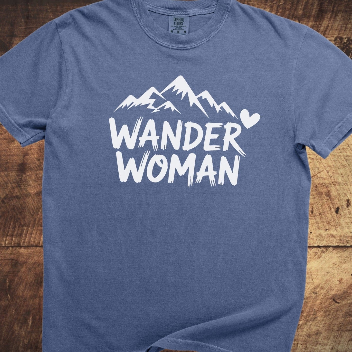 Wander Woman Comfort Colors 1717 Tee features a white mountain graphic and text on a medium-blue, short-sleeve shirt, offering a relaxed, durable fit.
