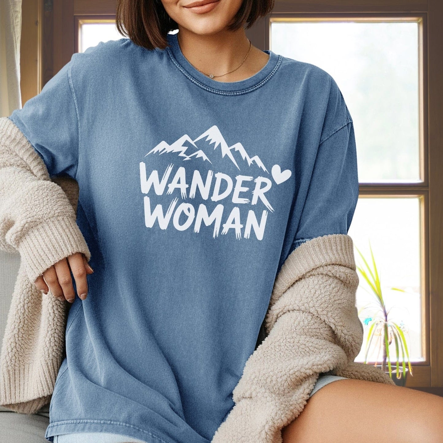 Person in a faded-blue Wander Woman Comfort Colors 1717 Tee with mountain graphic, draped cream sherpa cardigan, seated indoors near a window.