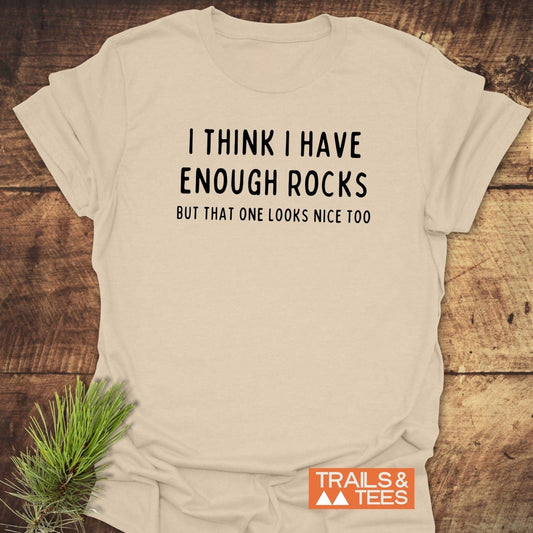 Enough Rocks T-Shirt on wood surface with pine sprig, bold black text, orange Trails & Tees logo. Soft ring-spun cotton, pre-shrunk, durable print.