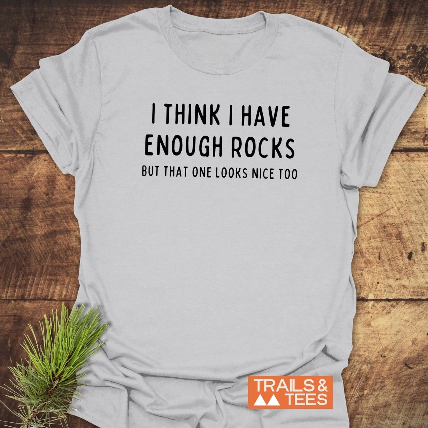Enough Rocks T-Shirt on wooden surface, features bold text about rocks. Includes TRAILS & TEES logo. Made from soft, durable, pre-shrunk cotton for outdoor enthusiasts.