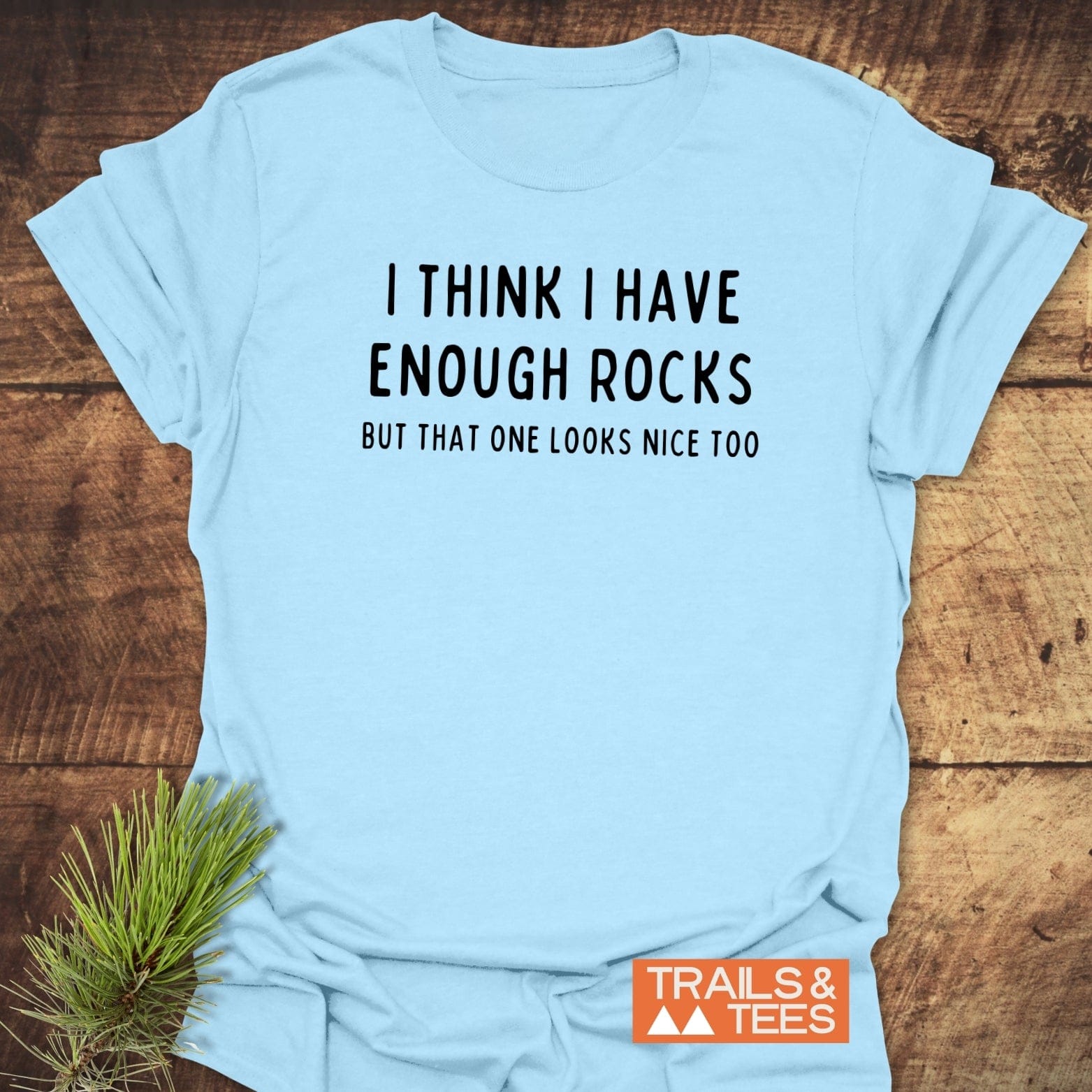Enough Rocks T-Shirt on rustic wood, featuring a playful slogan. Soft, lightweight cotton ideal for outdoor enthusiasts. Includes Trails & Tees logo and pine sprig accent.