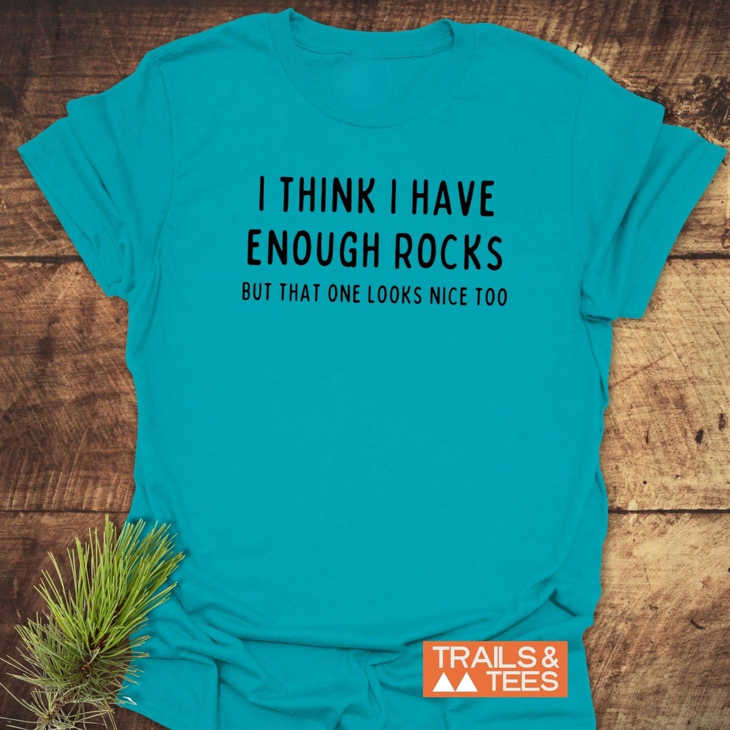 Enough Rocks T-Shirt with humorous outdoor-themed quote, laid flat with pine sprig accent. Features durable print and soft cotton for everyday adventures.