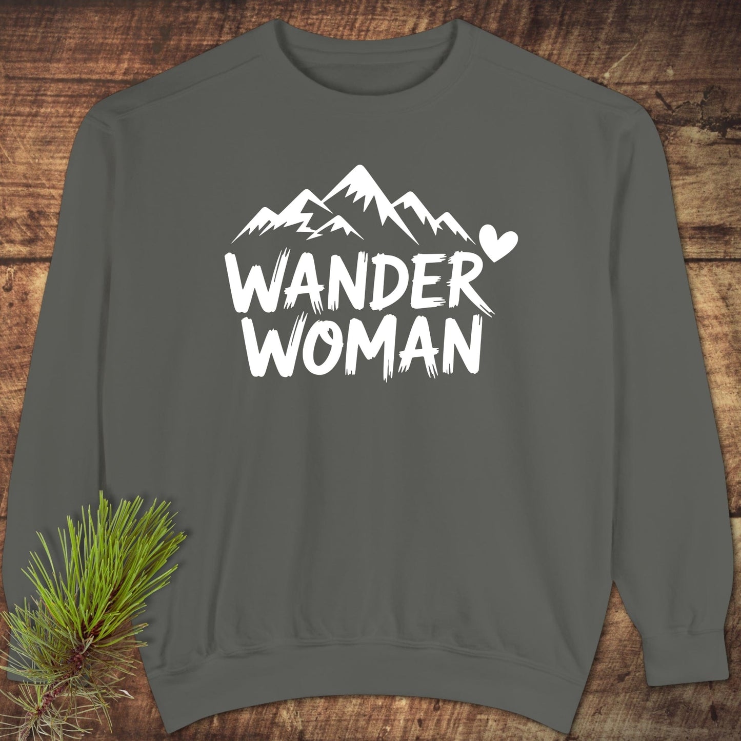 Wander Woman Sweatshirt with white mountain graphic, heart, and text on dark gray fabric, displayed on wood with pine sprig. Unisex, relaxed fit, garment-dyed fleece.