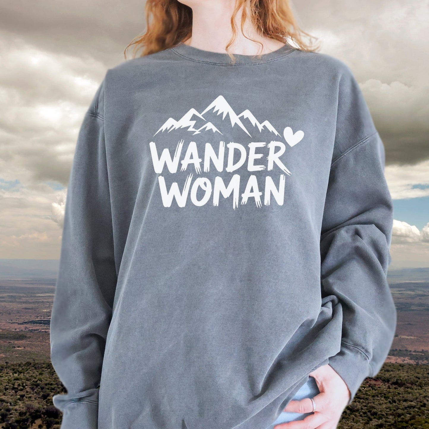 Person wearing the Wander Woman sweatshirt featuring a mountain graphic and heart. Oversized fit, slate-gray, made with ring-spun cotton and polyester blend.