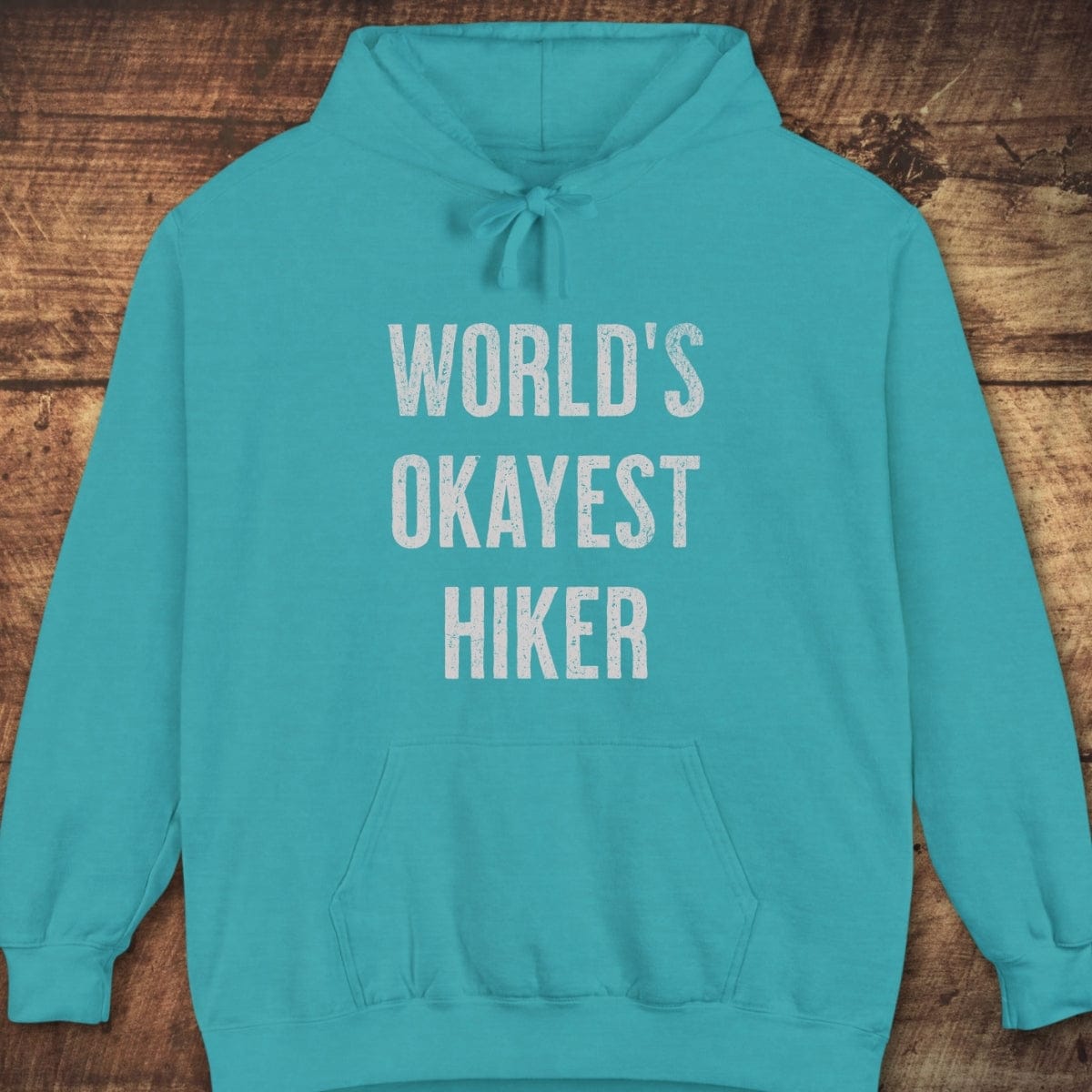 Teal hooded sweatshirt reads “WORLD’S OKAYEST HIKER” in white. Features a relaxed fit, jersey-lined hood, and front pouch pocket.