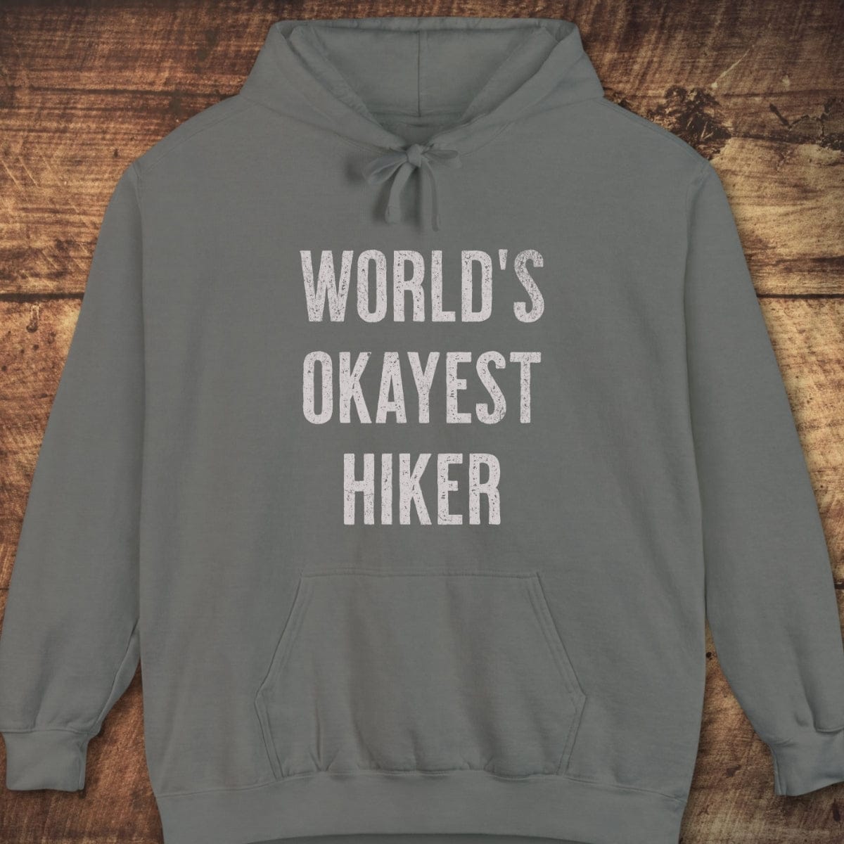 World's Okayest Hiker Comfort Colors Hoodie with distressed lettering, front pouch, jersey-lined hood, and relaxed fit, laid flat on a wooden surface.
