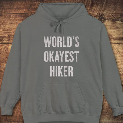 World's Okayest Hiker Comfort Colors Hoodie with distressed lettering, front pouch, jersey-lined hood, and relaxed fit, laid flat on a wooden surface.