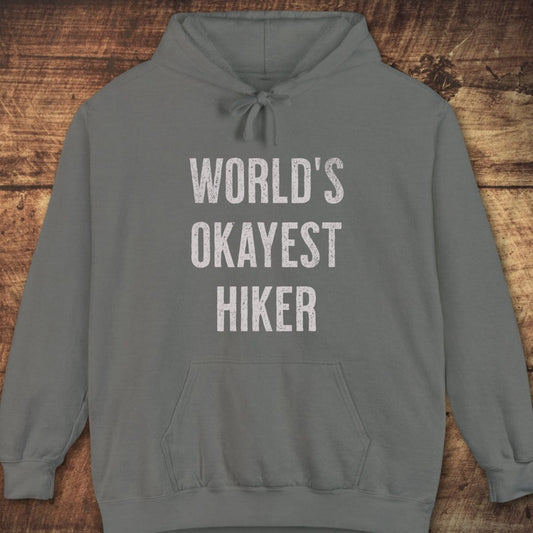 World's Okayest Hiker Comfort Colors Hoodie with distressed lettering, front pouch, jersey-lined hood, and relaxed fit, laid flat on a wooden surface.