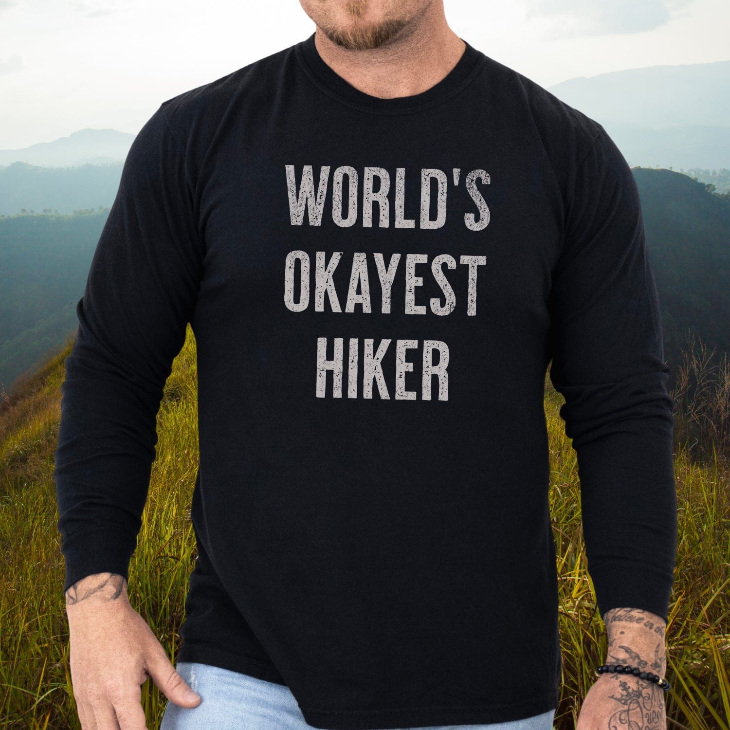 Person in hilly landscape wearing World's Okayest Hiker black long-sleeve tee, showcasing tattoos and bead bracelet, paired with light jeans.