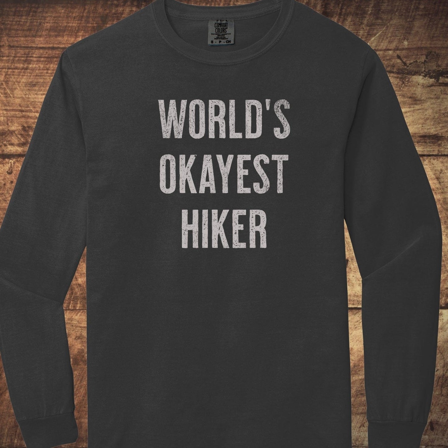 World's Okayest Hiker Comfort Colors Long-Sleeve Tee laid on wood, featuring distressed white lettering, 100% ring-spun cotton, relaxed fit, and garment-dyed fabric.