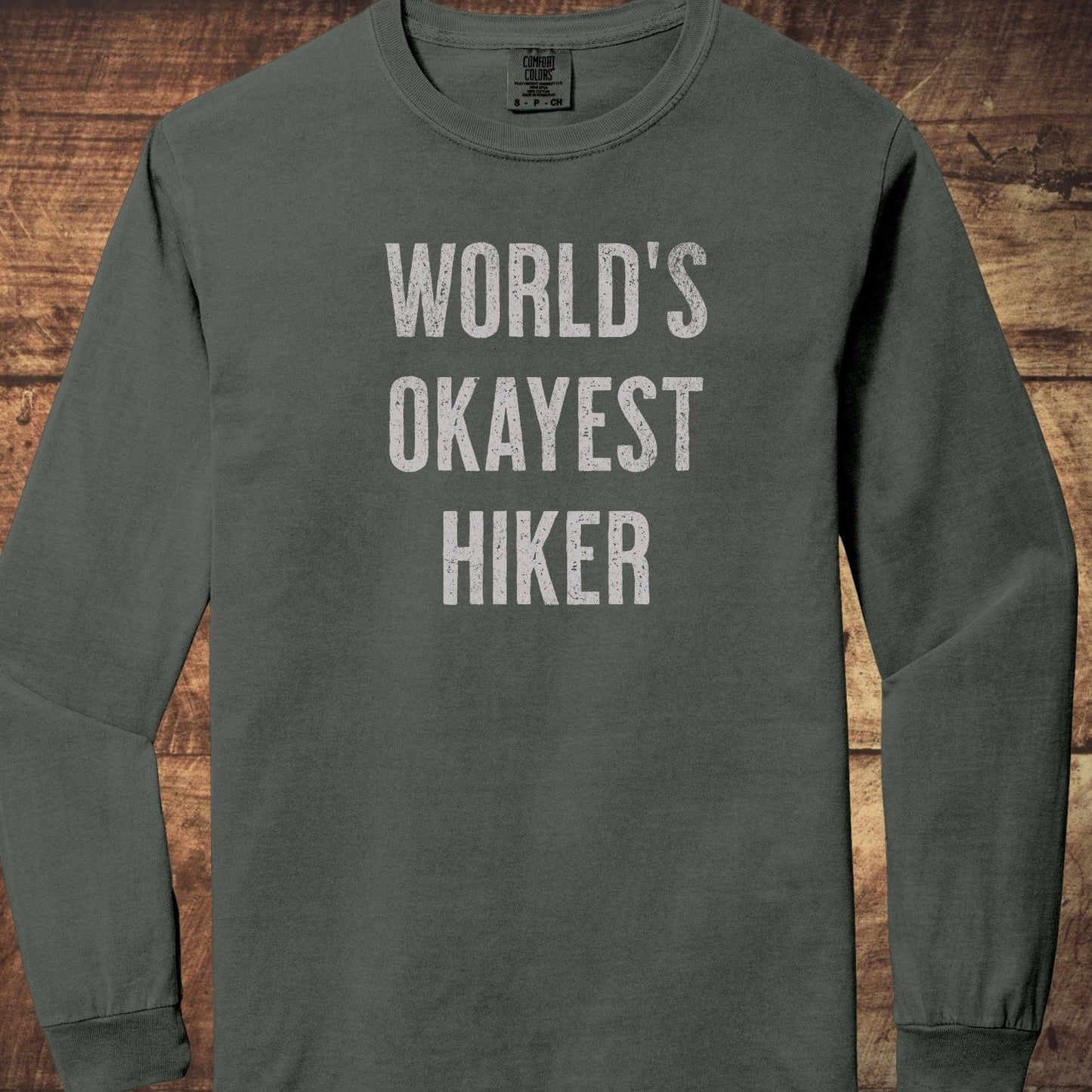 World's Okayest Hiker Comfort Colors Long-Sleeve Tee; features distressed lettering, laid flat, showcasing its relaxed, garment-dyed cotton fabric and classic fit.