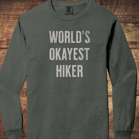 World's Okayest Hiker Comfort Colors Long-Sleeve Tee; features distressed lettering, laid flat, showcasing its relaxed, garment-dyed cotton fabric and classic fit.