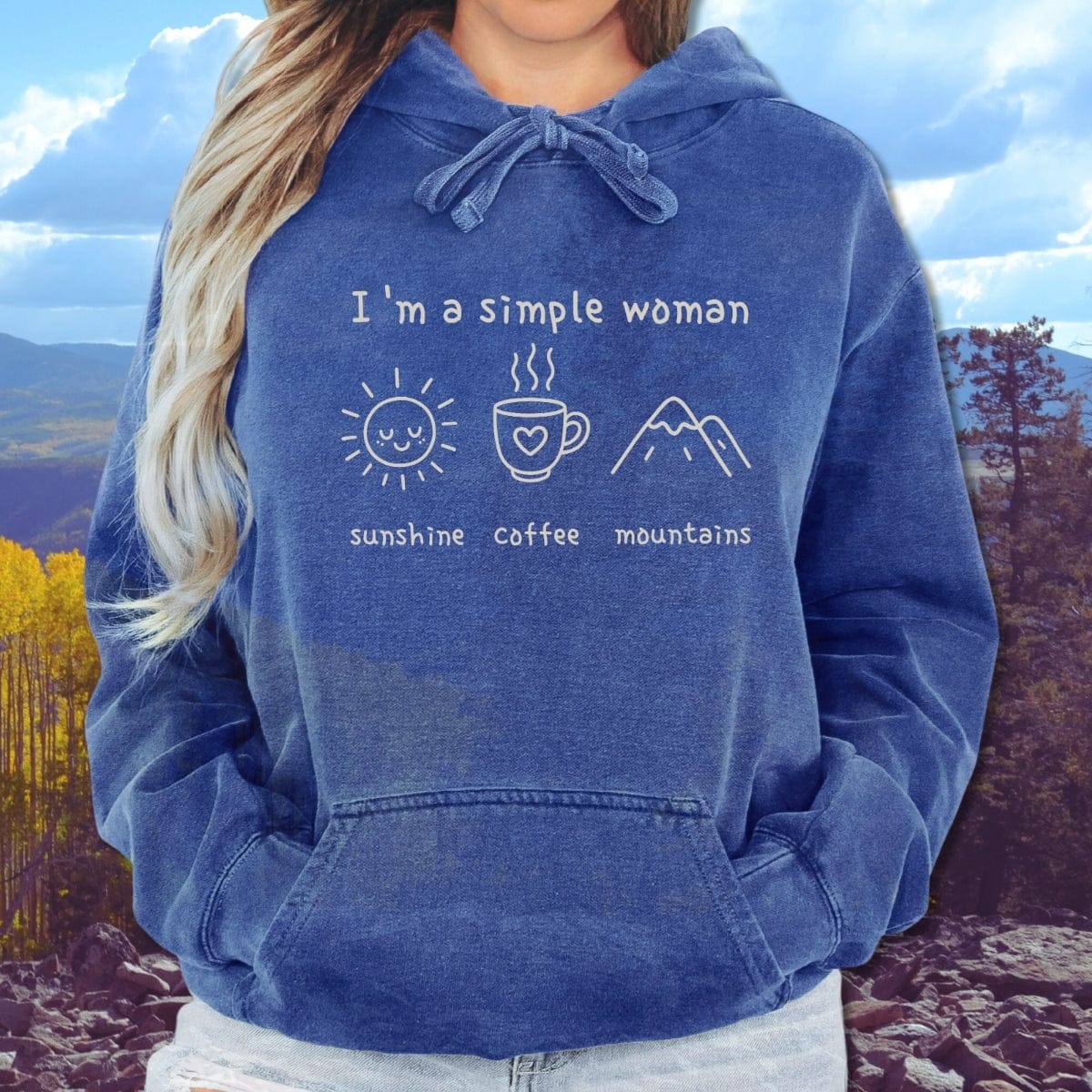Person wearing the I'm A Simple Woman Comfort Colors Hoodie featuring icons of a sun, coffee mug, and mountains, set against a forested mountain backdrop.