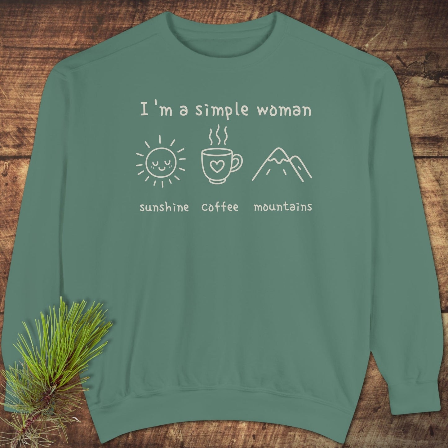 I'm A Simple Woman Comfort Colors 1566 Sweatshirt with 'sunshine, coffee, mountains' icons on teal-green fabric, laid on wooden surface with pine sprig.