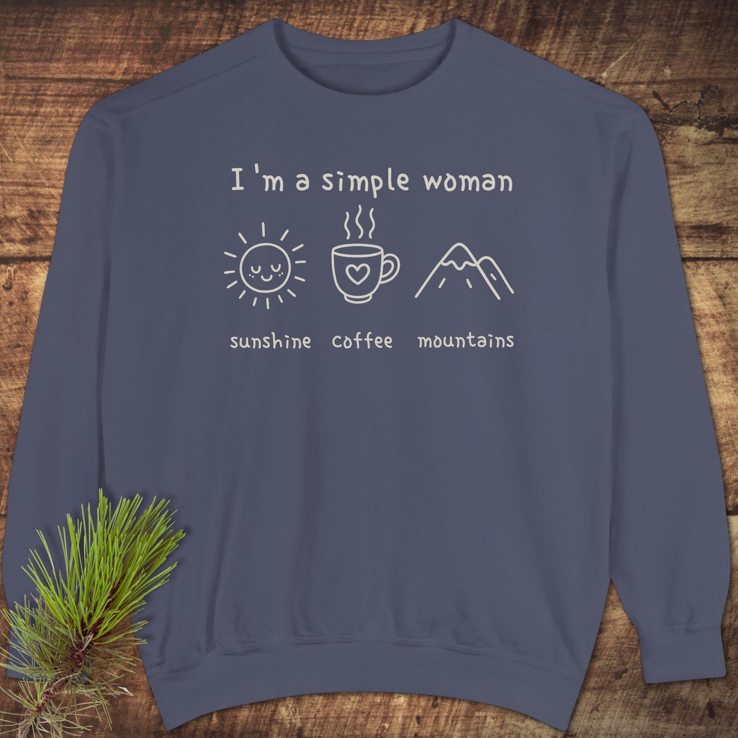 I'm A Simple Woman Comfort Colors 1566 Sweatshirt with 'sunshine, coffee, mountains' graphic, laid flat on wood, accompanied by a pine sprig.