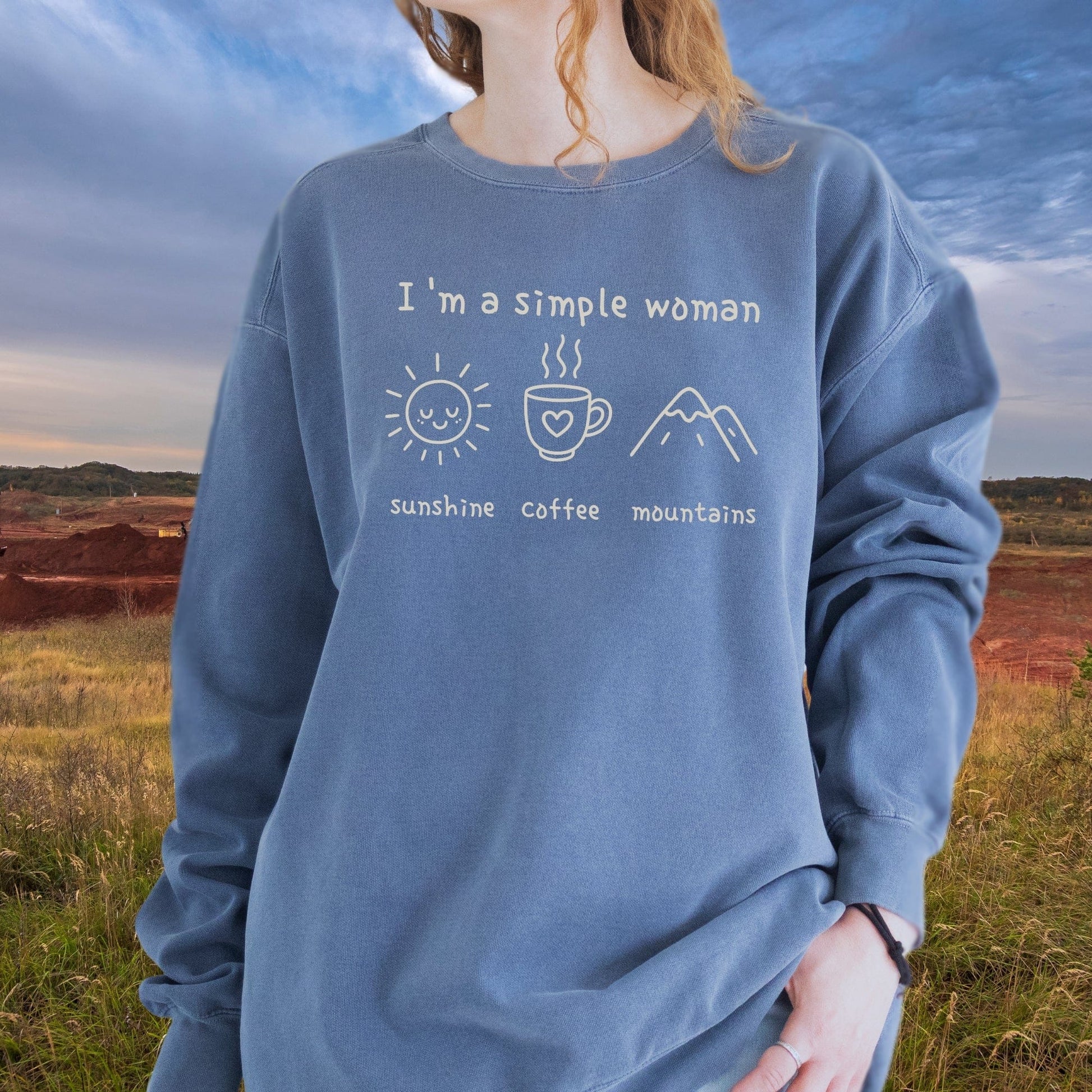 Person wearing the I'm A Simple Woman Comfort Colors 1566 Sweatshirt featuring icons for sunshine, coffee, and mountains, standing outdoors in a relaxed fit garment.
