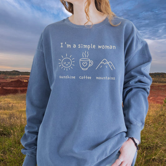 Person wearing the I'm A Simple Woman Comfort Colors 1566 Sweatshirt featuring icons for sunshine, coffee, and mountains, standing outdoors in a relaxed fit garment.