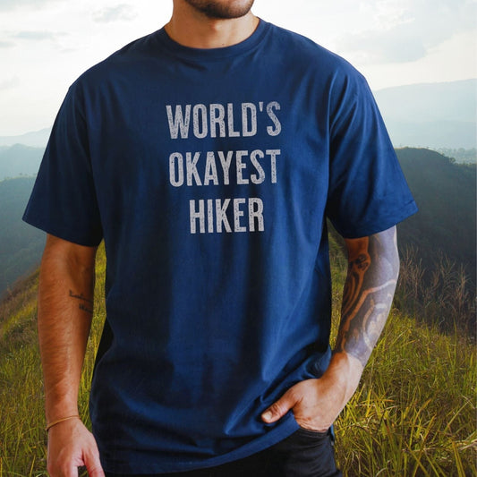 Man wearing World's Okayest Hiker Comfort Colors 1717 Tee, showcasing its relaxed fit and soft-washed texture, standing on a grassy hillside.