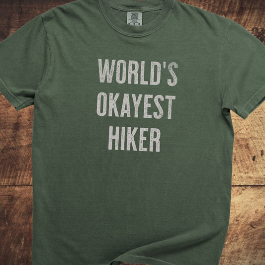 World's Okayest Hiker Comfort Colors 1717 Tee in forest-green, short-sleeve, with distressed white lettering, displayed flat on a wooden surface, showcasing its relaxed fit.