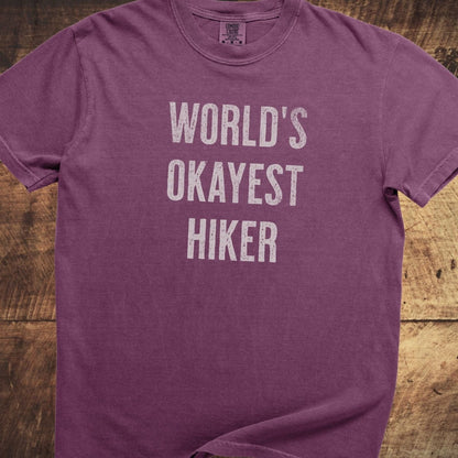 World's Okayest Hiker Comfort Colors 1717 Tee in purple heathered fabric, featuring distressed white text, soft-washed cotton, relaxed fit, laid on wood surface.