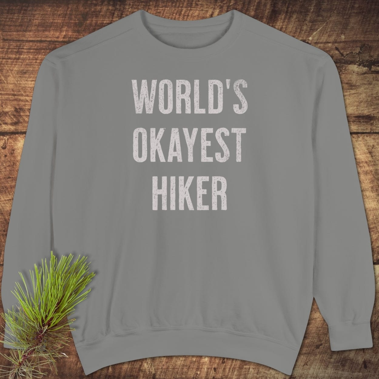 Light-grey World's Okayest Hiker sweatshirt with distressed white lettering, shown flat with pine bough. Features relaxed fit and garment-dyed cotton-polyester blend.