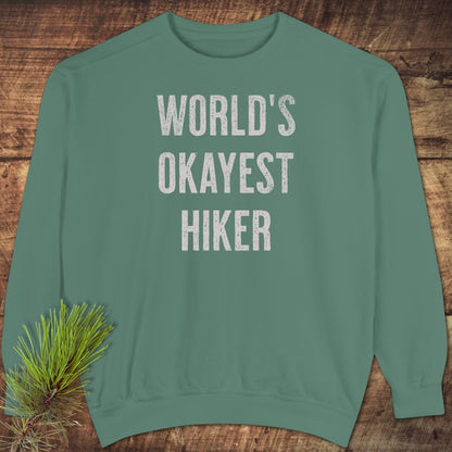 World's Okayest Hiker Comfort Colors Sweatshirt, green with white slogan, laid flat on wood; includes relaxed fit, garment-dyed fabric, pine sprig accent.