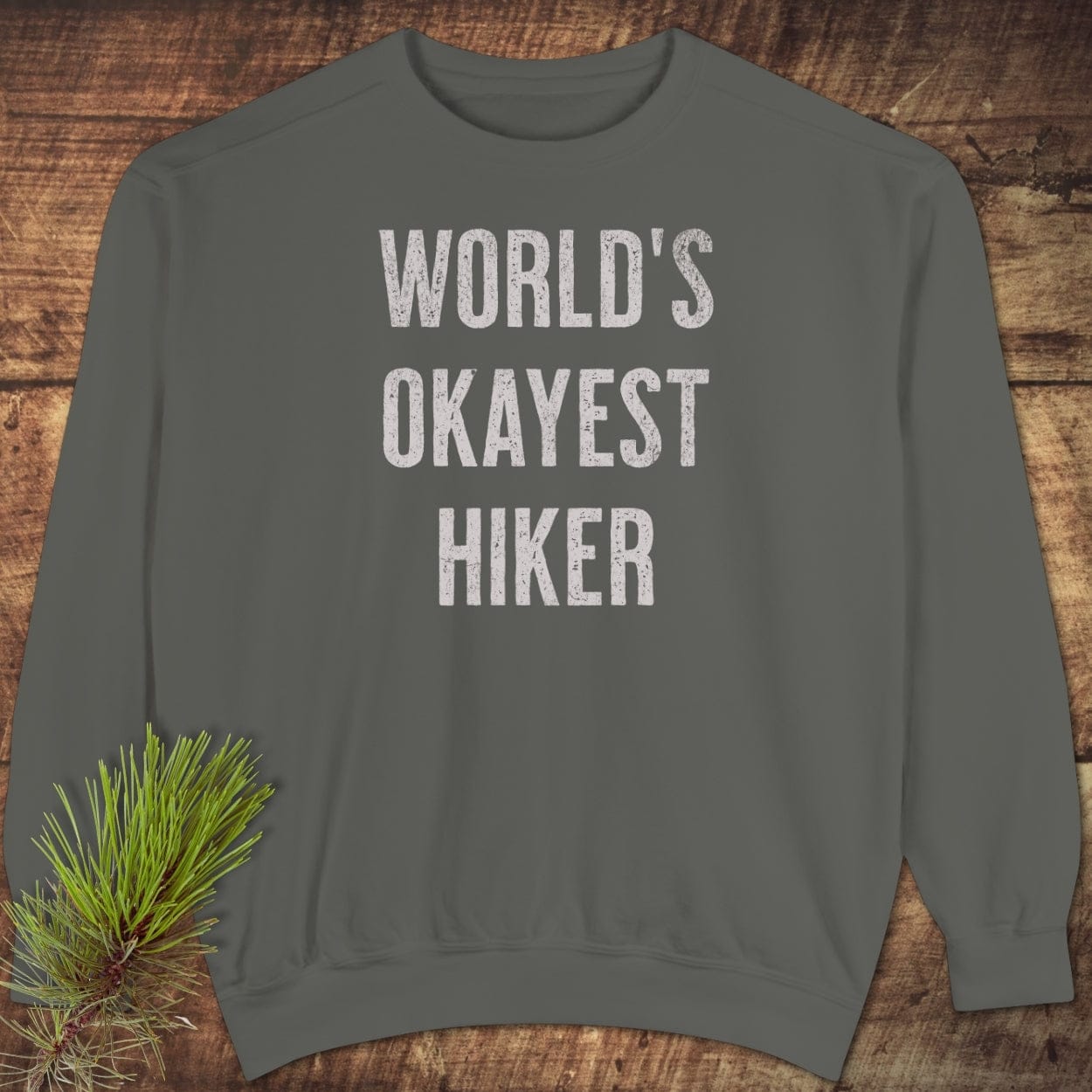 World's Okayest Hiker Comfort Colors Sweatshirt, dark-olive, crewneck, long-sleeve, laid flat with rustic pine branch, showcasing relaxed fit and weathered white print.