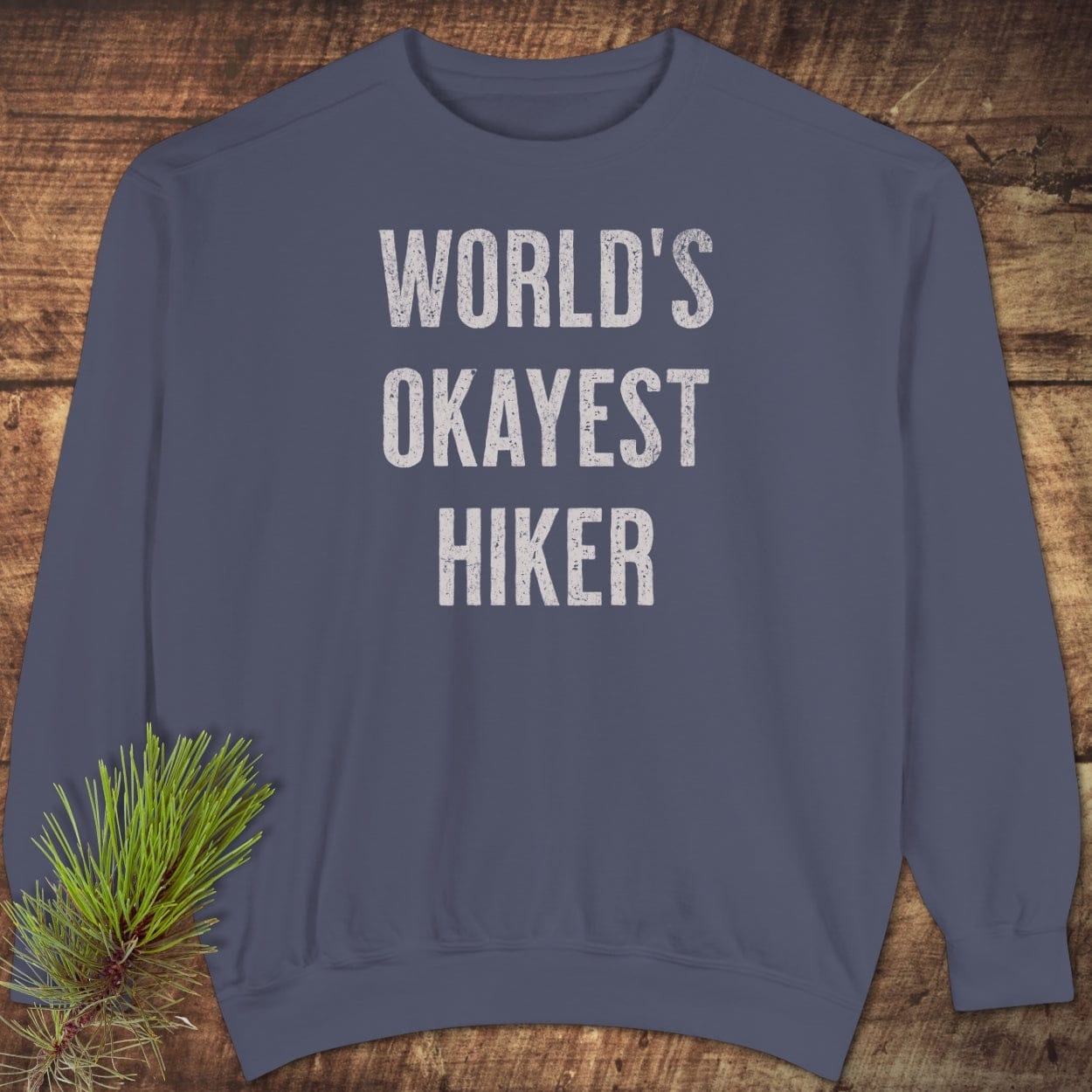 Slate-blue crewneck sweatshirt with 'WORLD’S OKAYEST HIKER' text, showcasing relaxed fit and garment-dyed fleece, laid on a wooden surface with pine boughs.