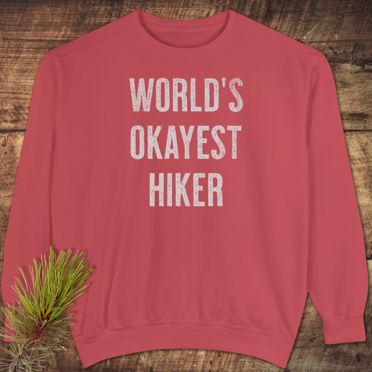 Red World’s Okayest Hiker sweatshirt on wood surface, featuring bold white lettering, relaxed fit, and pine sprig accentuating its outdoorsy appeal.