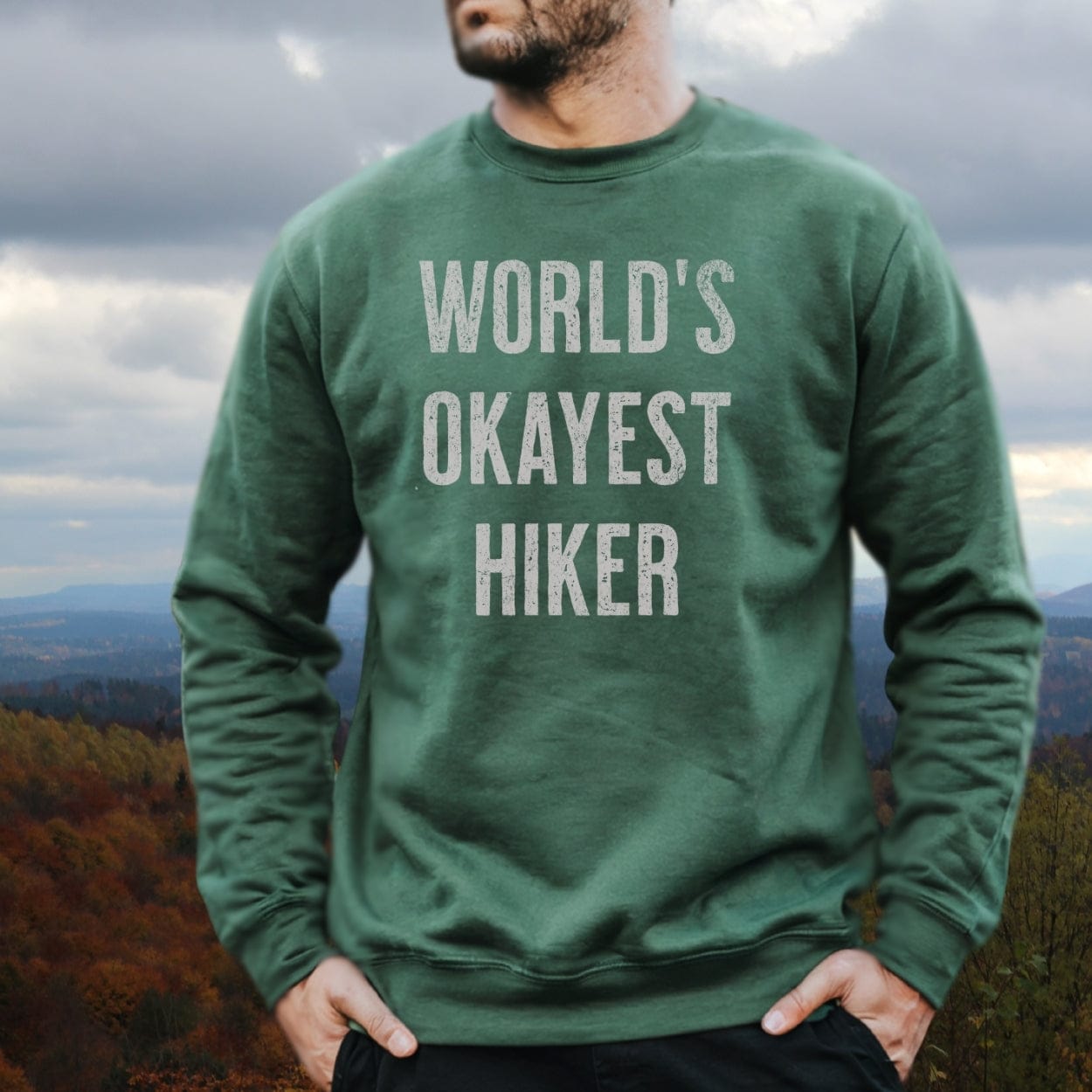 Person outdoors wearing a WORLD’S OKAYEST HIKER green sweatshirt, highlighting its relaxed fit and luxurious comfort from the World's Okayest Hiker Comfort Colors Sweatshirt line.