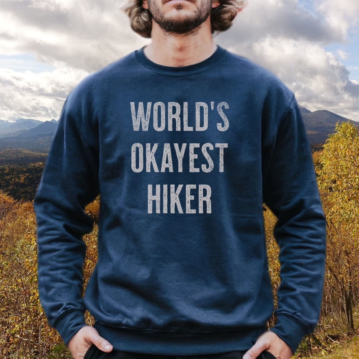Person outdoors wearing World's Okayest Hiker navy sweatshirt, showcasing relaxed fit and comfort with luxurious garment-dyed fabric. Autumnal forest backdrop visible.
