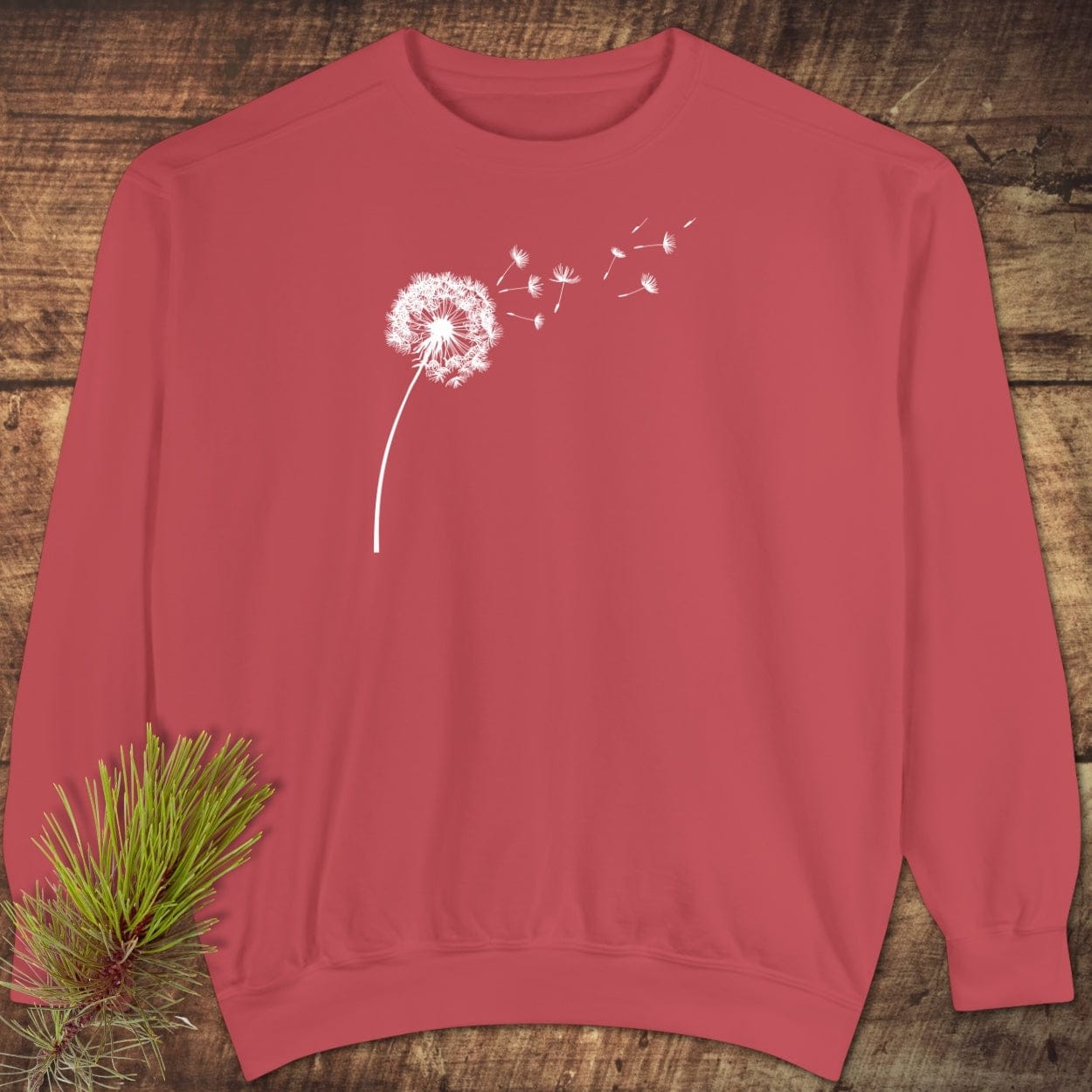 Dandelion Comfort Colors Sweatshirt on wood surface; features white dandelion print, relaxed fit, 80% cotton, 20% polyester. Pine sprig nearby.